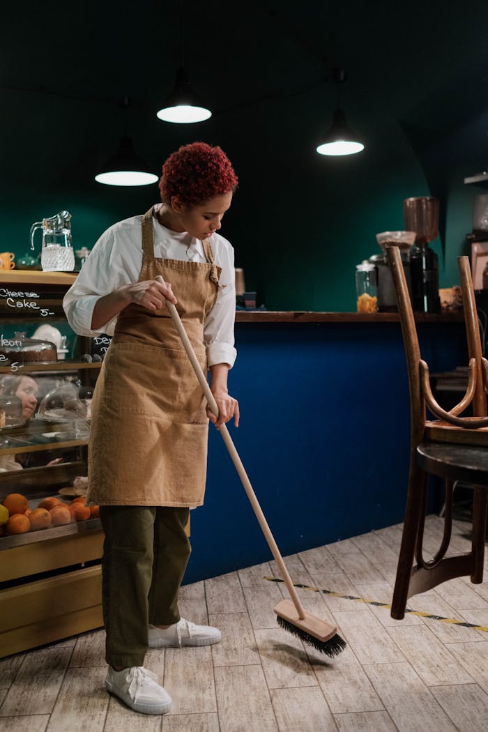 A barista in an apron sweeps the café floor, maintaining cleanliness in a cozy indoor setting.